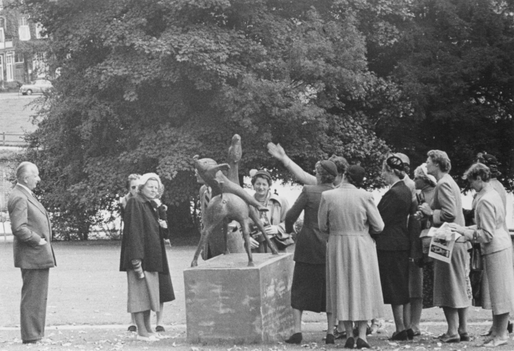Op deze zwart-wit foto wordt een bronzen beeld op een sokkel omgeven door een groep van elf vrouwen die dicht bij het beeld staan. Aan de linkerkant staat koningin Juliana, met links van haar en man. Het bronzen beeld is van achteren gefotografeerd. Het is een ruiter te paard. De ruiter leunt heel ver naar achteren. Het paard houdt het hoofd verticaal omhoog. Een van de vrouwen houdt haar arm schuin omhoog. Ze lijkt de beweging van de ruiter van het beeld te kopiëren. Het gezelschap en het beeld bevinden zich in Park Sonsbeek, op een grasveld omgeven door grote bomen.