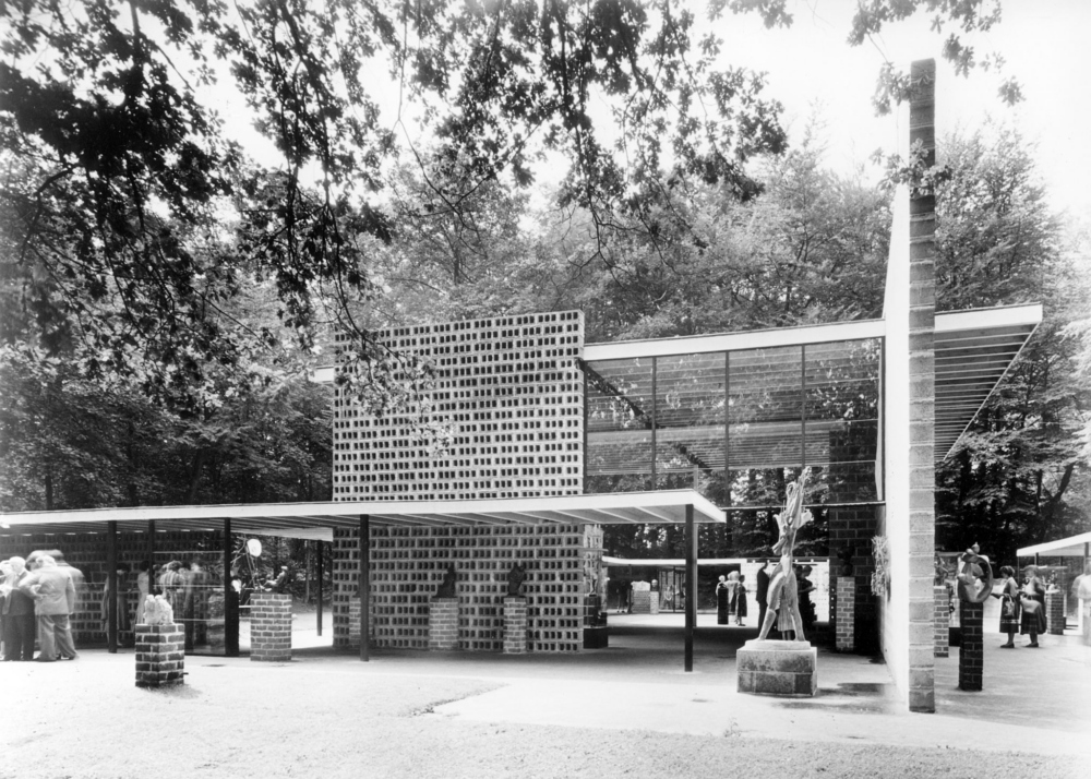 Op deze zwart-wit foto wordt de aandacht getrokken door een groot geometrisch paviljoen van zeker twintig meter breed met open structuur. Het paviljoen wordt omgeven door hoge bomen, vol in het blad. Binnenin en rondom het paviljoen staan beeldhouwwerken. Verder lopen er bezoekers rond.