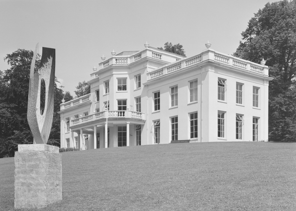 De Witte Stadsvilla van Park Sonsbeek is prominent in beeld op deze zwart-wit foto. Aan de linkerkant, op de voorgrond, staat een kunstwerk op een sokkel van ongeveer een meter hoog. Het is een verticale, langwerpig gevormde sculptuur met een gat in het midden. Metalen draden zijn vanaf de randen over de sculptuur gespannen, alsof een spin er een web op heeft gemaakt.