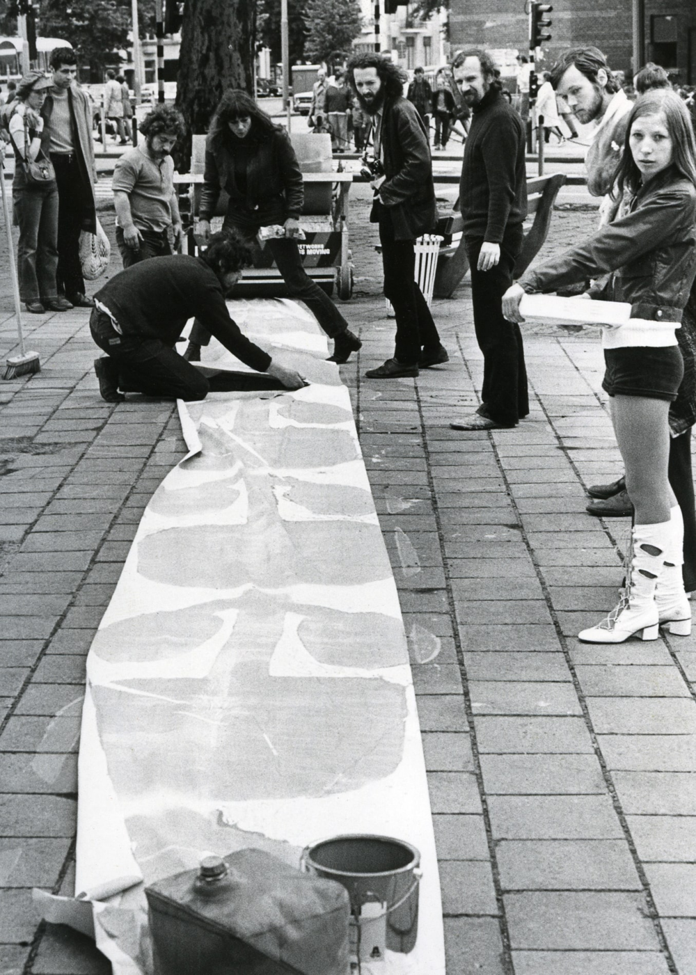 Een zwart-wit foto die op een drukke stoep in de stad gemaakt lijkt te zijn. In de achtergrond zijn mensen te zien en een stoplicht. Een smal wit vel papier met bloemmotief van zo’n vijf meter lang ligt op de stoep. Een zwart geklede man zit gehurkt op zijn knieën op het vel papier met een onbekend instrument. Drie andere zwart geklede mannen stan vlak bij hem en kijken mee. Rechts in beeld staat een jonge vrouw met opvallende witte laarzen met blokhakken.