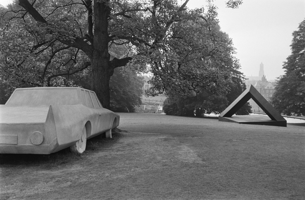 Een zwart-wit foto van twee verschillende kunstwerken op een grasheuvel in Park Sonsbeek. Links staat een stenen kunstwerken van een auto tegen een grote boom aan. Het is alsof de auto tegen de boom is gebotst. De auto is gestileerd weergegeven. Er zijn weinig specifieke kenmerken, zoals een logo van een merk. Wat lager geleden op de heuvel, aan de rechterkant, staat een groot stalen kunstwerk. Het bestaat uit een vierkante, vlakke bodem met een driehoek als een soort poort. Je zou onder het werk door kunnen lopen.