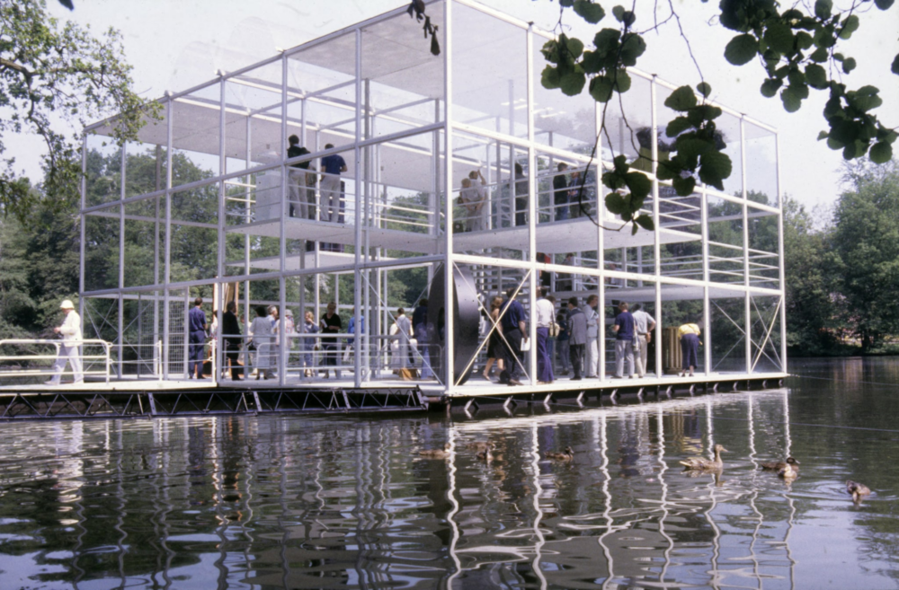 Op een grote vijver in park Sonsbeek drijft een groot kubus gevormd paviljoen van glas. Het heeft een verdieping waar mensen rondlopen. Op de begane grond staan een aantal kunstwerken met veel mensen eromheen. Bij de ingang van het paviljoen aan de voorkant ligt een loopbrug op het water die het paviljoen ogenschijnlijk met het land verbindt.