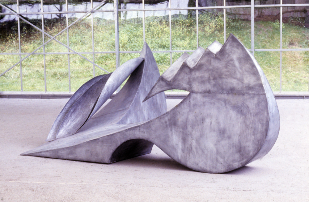 Deze foto is van een abstract kunstwerk van metaal dat op een stenen vloer ligt. Het kunstwerk is grijs gemeleerd en glimt niet. Het bevat ronde en hoekige, trapachtige vormen. Het werk lijkt in een soort kas te liggen, want op de achtergrond van de foto is een ijzeren structuur met allerlei ramen afgebeeld.