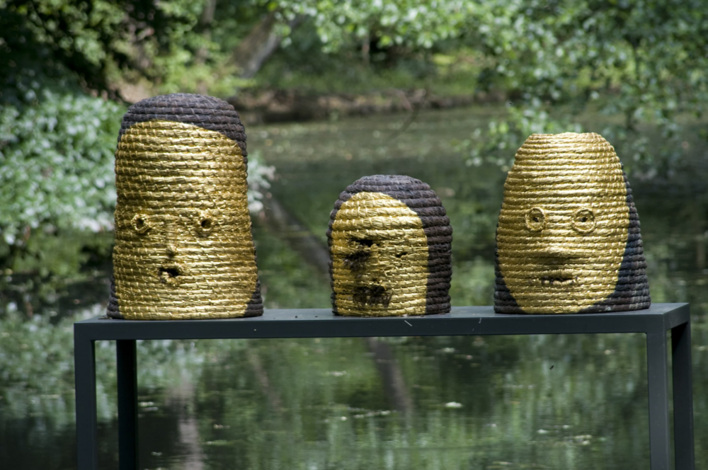 Three beehives in the shape of heads. The mouths and eyes of the figures are openings for the bees. The hives are woven from straw and mostly gold-colored. They stand on a black table in a small pond, surrounded by trees in Sonsbeek Park.