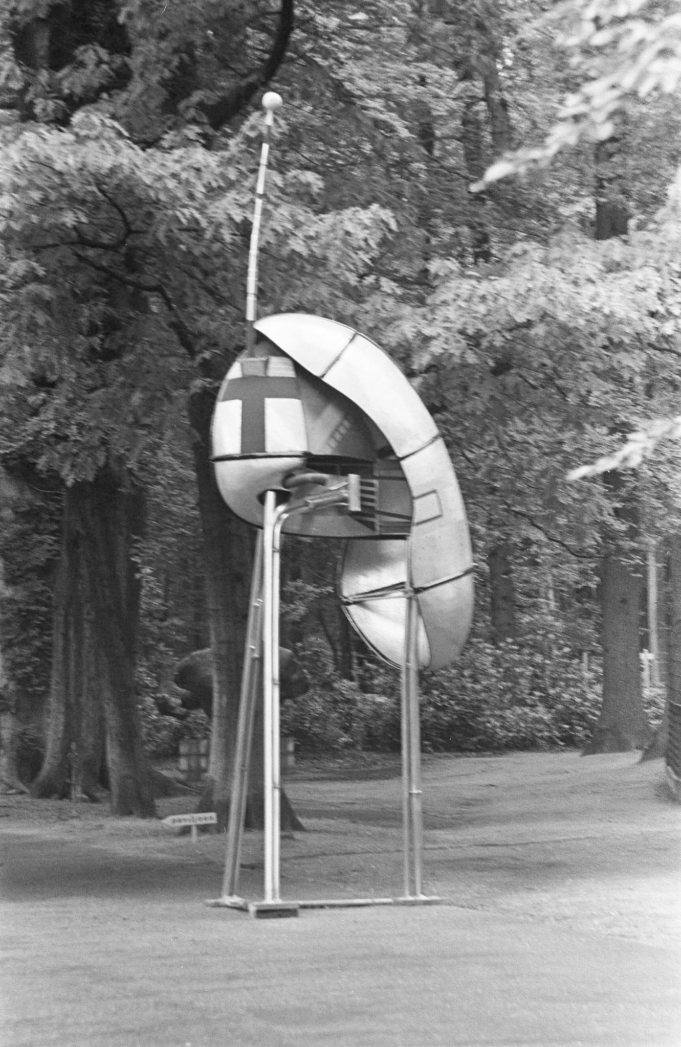 Op deze zwart-wit foto staat een merkwaardig object. Het doet denken aan een zendmast, vanwege de drie poten en een soort antenne. Maar het is vele malen kleiner dan een daadwerkelijke zendmast. Het heeft ook iets weg van een ruimteschip dat net geland is. Het staat op het gras, omgeven door hoge bomen vol in het blad.