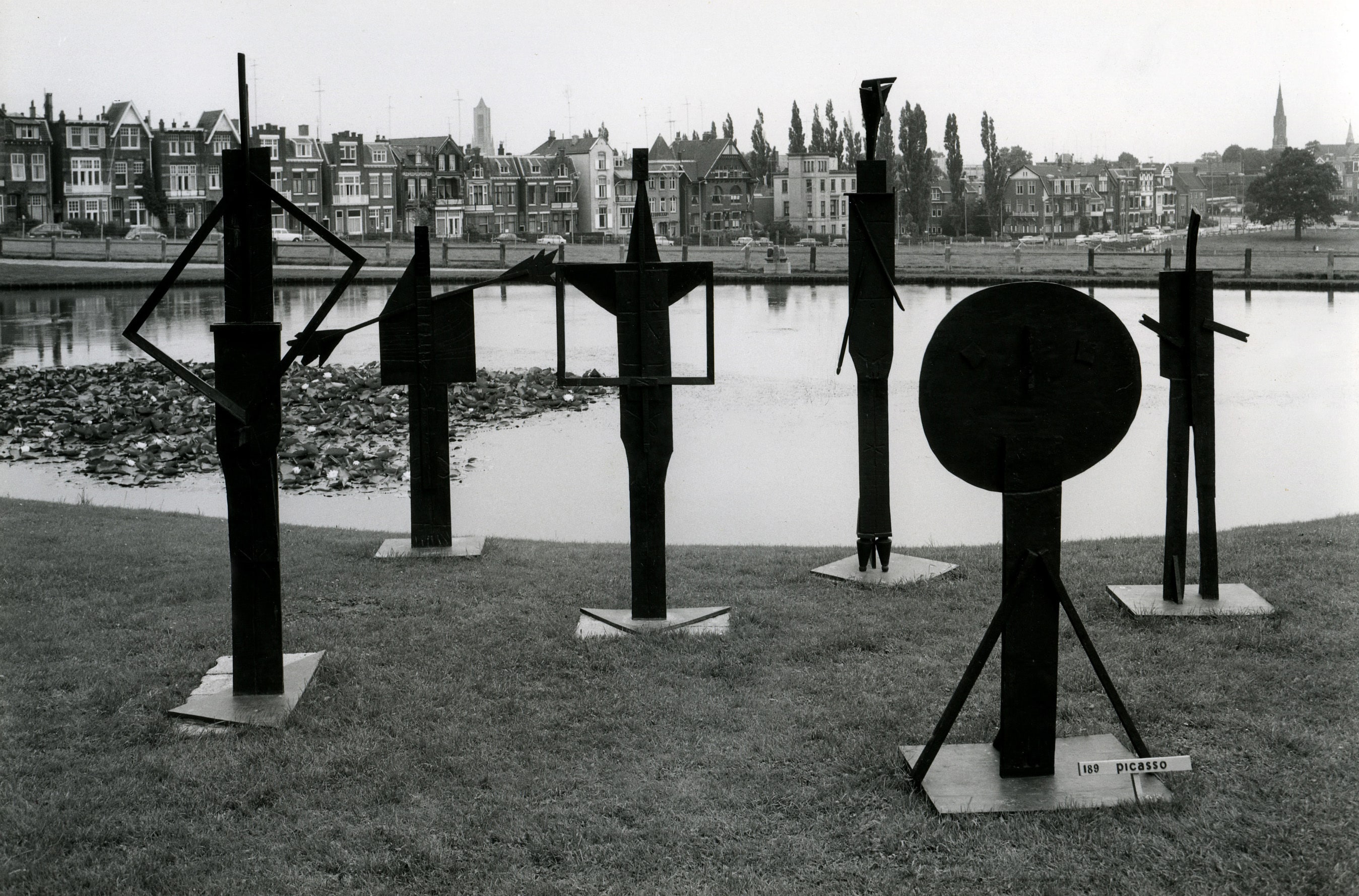 Op deze zwart-wit foto staat een beeldengroep van zes zwart uitziende beelden op het gras, zonder sokkel. Ze staan dicht bij elkaar, hoewel je wel om de beelden heen zou kunnen lopen. Ze staan pal aan een grote vijver. Het zijn abstracte, verticale, smalle beelden. Ze staan op kleine vierkante plateaus, om ze overeind te houden. Bij het beeld dat het meest op de voorgrond staat, staat een bordje waarop de naam Picasso staat geschreven. Op de achtergrond van de foto zijn de Sonsbeeksingel, de toren van de Eusebiuskerk en de binnenstad van Arnhem te herkennen.