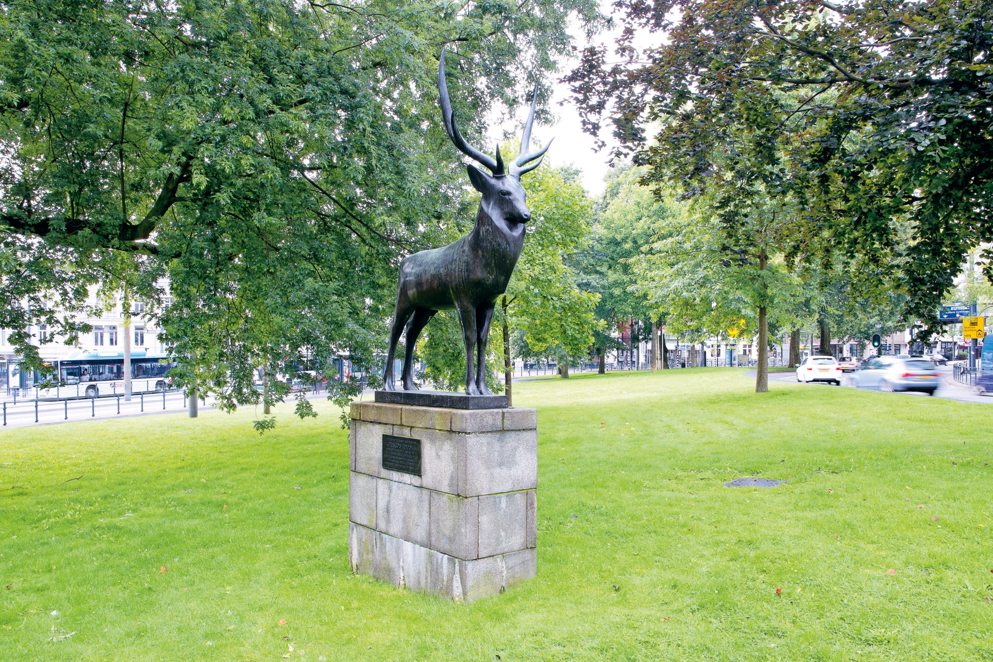 A bronze sculpture of a stag with antlers stands on a tall pedestal made of stone blocks, about one and a half meters high. The sculpture has a very smooth surface. Several large trees surround the sculpture.