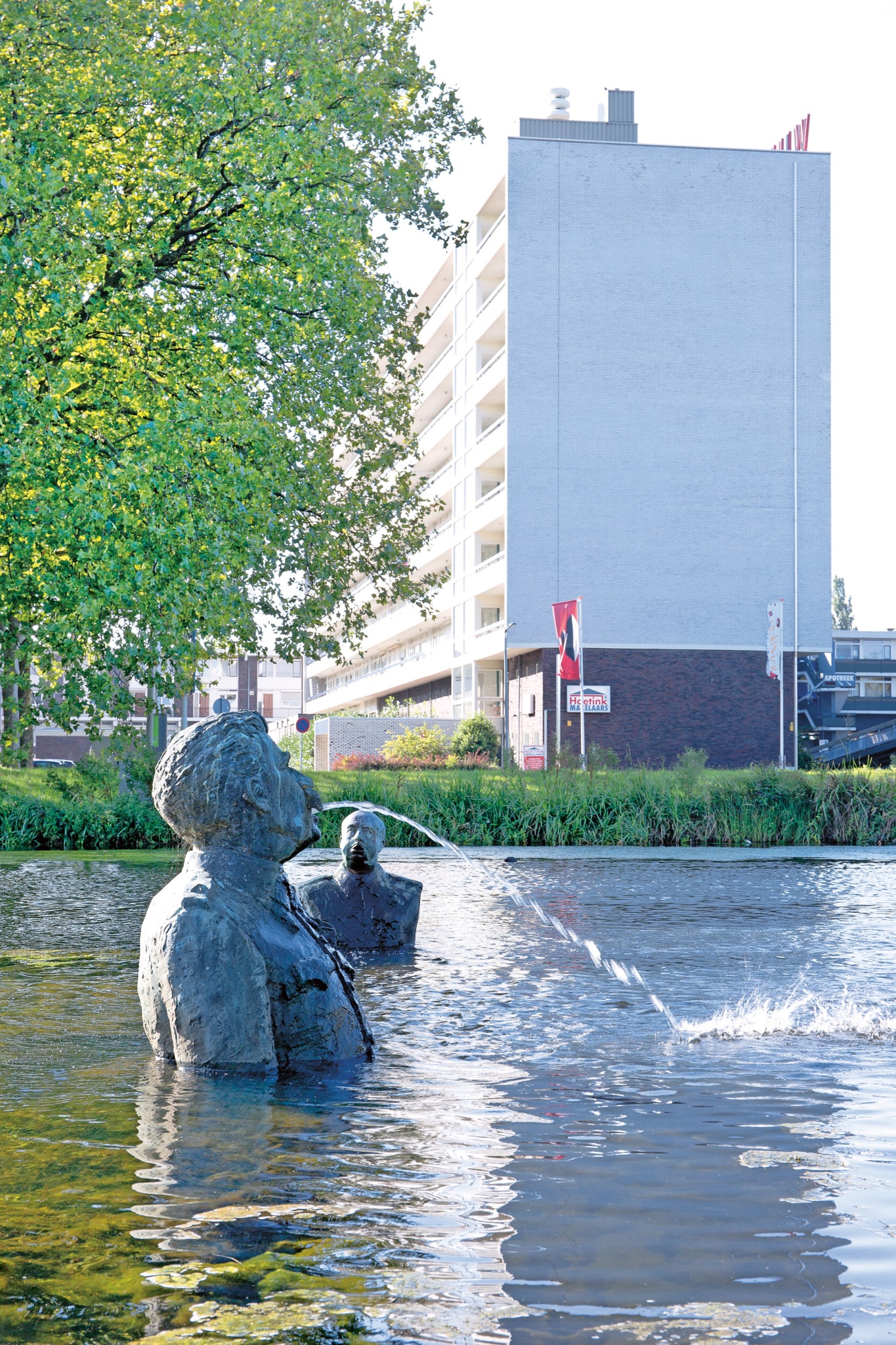 In a pond, a fountain is visible, consisting of bronze busts emerging from the water. The busts depict male figures, with the large man with a mustache in the foreground spouting a jet of water from his mouth. In the background stands a tall gallery apartment building, with a large, leafy tree on the left.