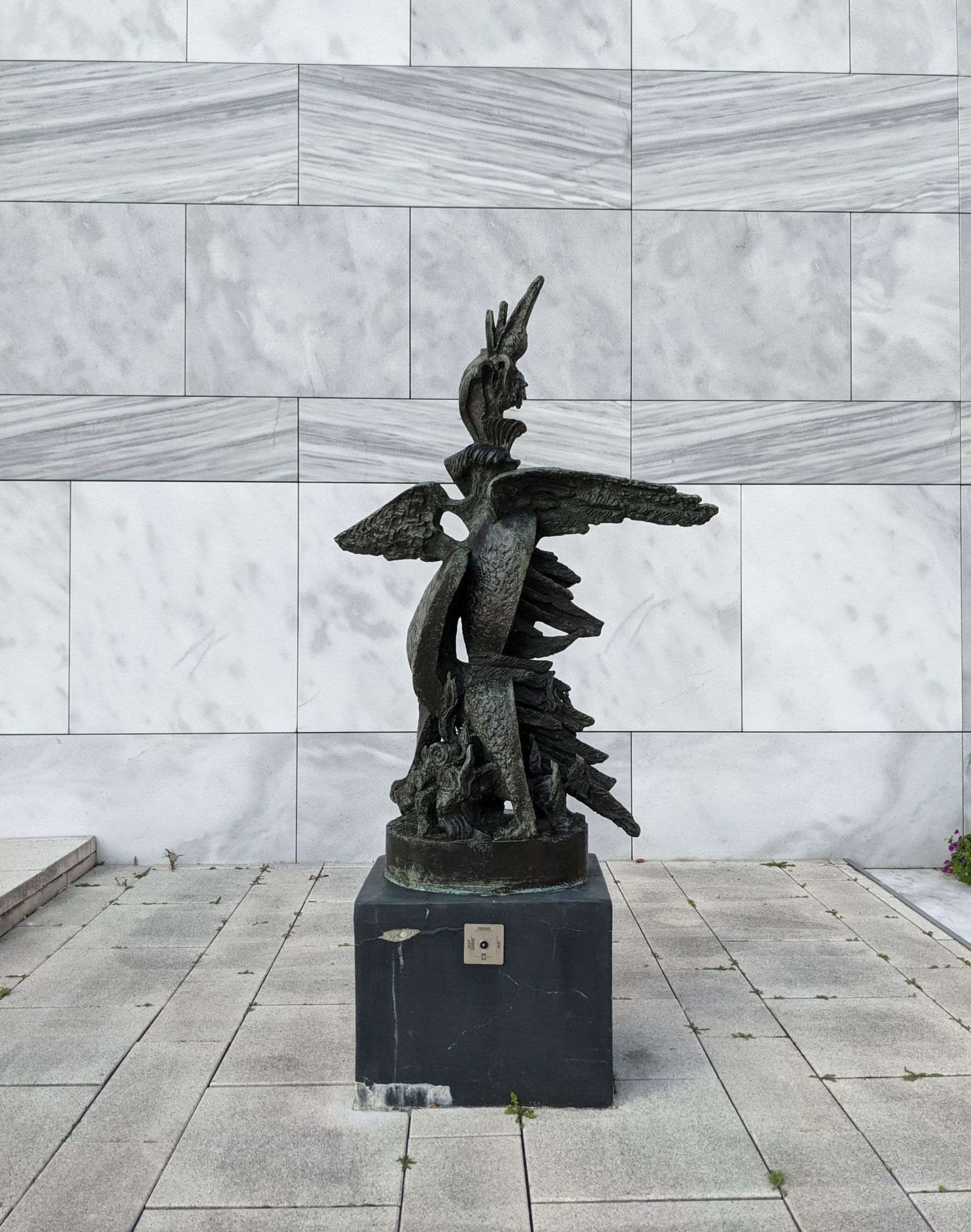 An abstract-looking bronze statue nearly two meters high stands on a square black pedestal about fifty centimeters tall. The sculpture contains recognizable elements of a bird: two outstretched wings, large protruding feathers, and at the top a bird’s head with an open beak pointing upward. It is as if the bird is calling for help. The statue stands right next to a grey marble wall.