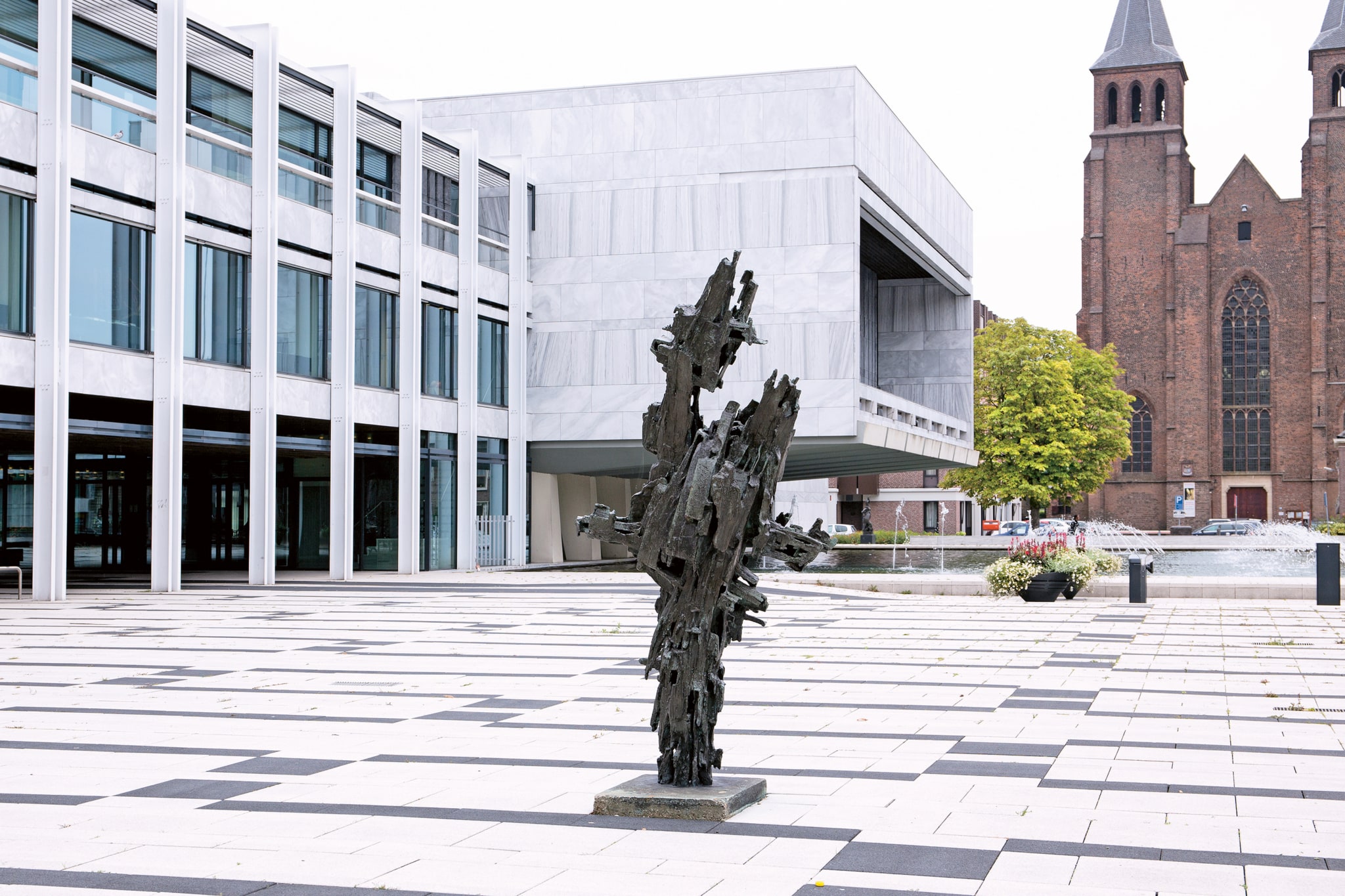 In a square with a large office-like building with marble walls, there is a bronze statue on a very low pedestal of about ten centimeters. It is an abstract sculpture with a vertical shape, featuring geometric cutouts and protruding parts. In the background is a pond with fountains and the front view of a brown church building.