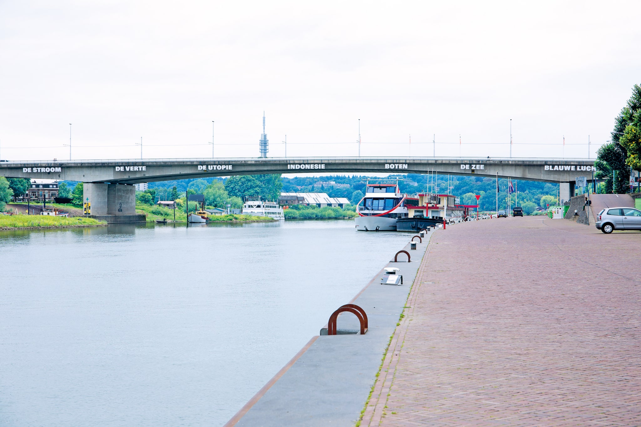 Een autobrug over een rivier met aan de rechterkant een stenen kade. Onder de brug liggen twee boten naast elkaar. Aan de zijkant van de brug zijn grote witte woorden in kapitalen te zien: van links naar rechts zijn dat de woorden de stroom, de verte, de utopie, Indonesië, boten, de zee, blauwe lucht.