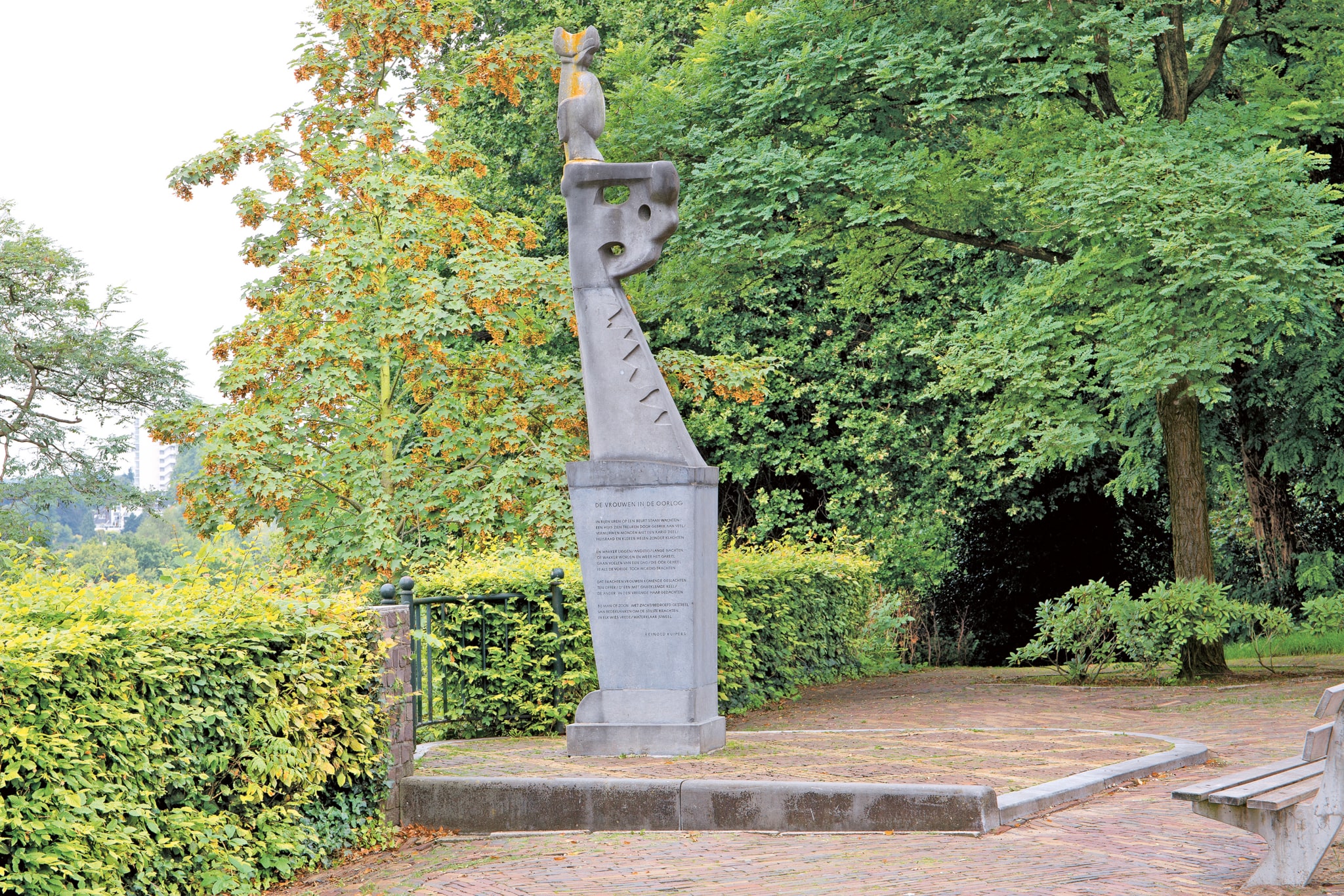 A stone statue stands on a man-high stone pedestal. There is an inscription on the pedestal. The small face reveals that the statue is of a female figure. Otherwise, the work is abstract; she appears to be carrying something on her head. She seems winged, like a bird. The statue stands along a low beech hedge full of leaves. To the right of the statue is a wooden bench.