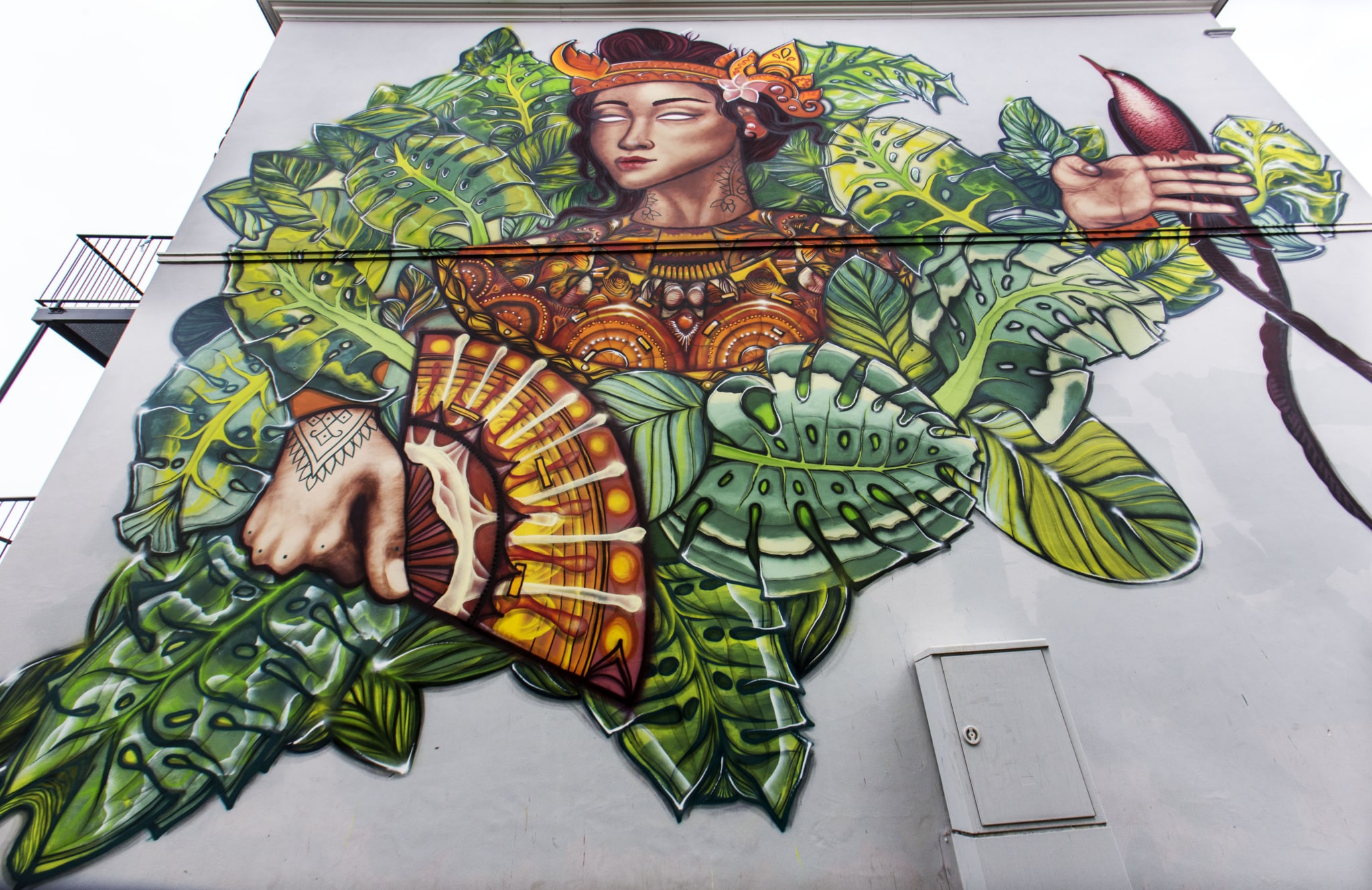 On the right side of a building at least three stories high, there is a large mural. This mural shows the face of a woman. She has a sort of collar and long dark green hair. She has two pairs of eyes, one directly below the other. The open eyes have no pupils. There are also floral motifs, with a large hand in the center. The index and middle fingers are raised.