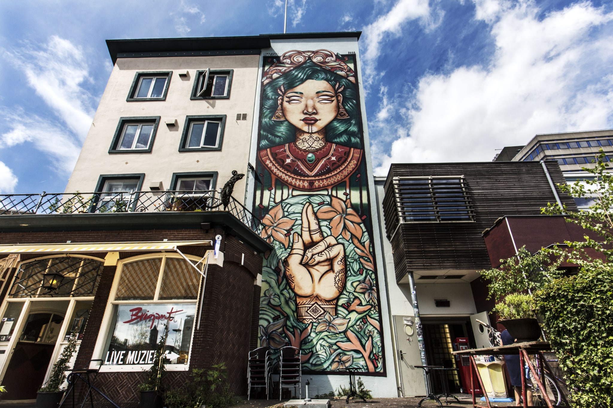 Op de rechterkant van een gebouw van ten minste drie verdiepingen hoog is een grote muurschildering te zien. Deze muurschildering bevat het gezicht van een vrouw. Ze heeft een soort kraag en donkergroen lang haar. Ze heeft twee paar ogen vlak onder elkaar. De geopende ogen bevatten geen pupillen. Verder zijn er bloemmotieven, met in het midden een grote hand. De wijs -en middelvinger zijn omhoog gestoken.