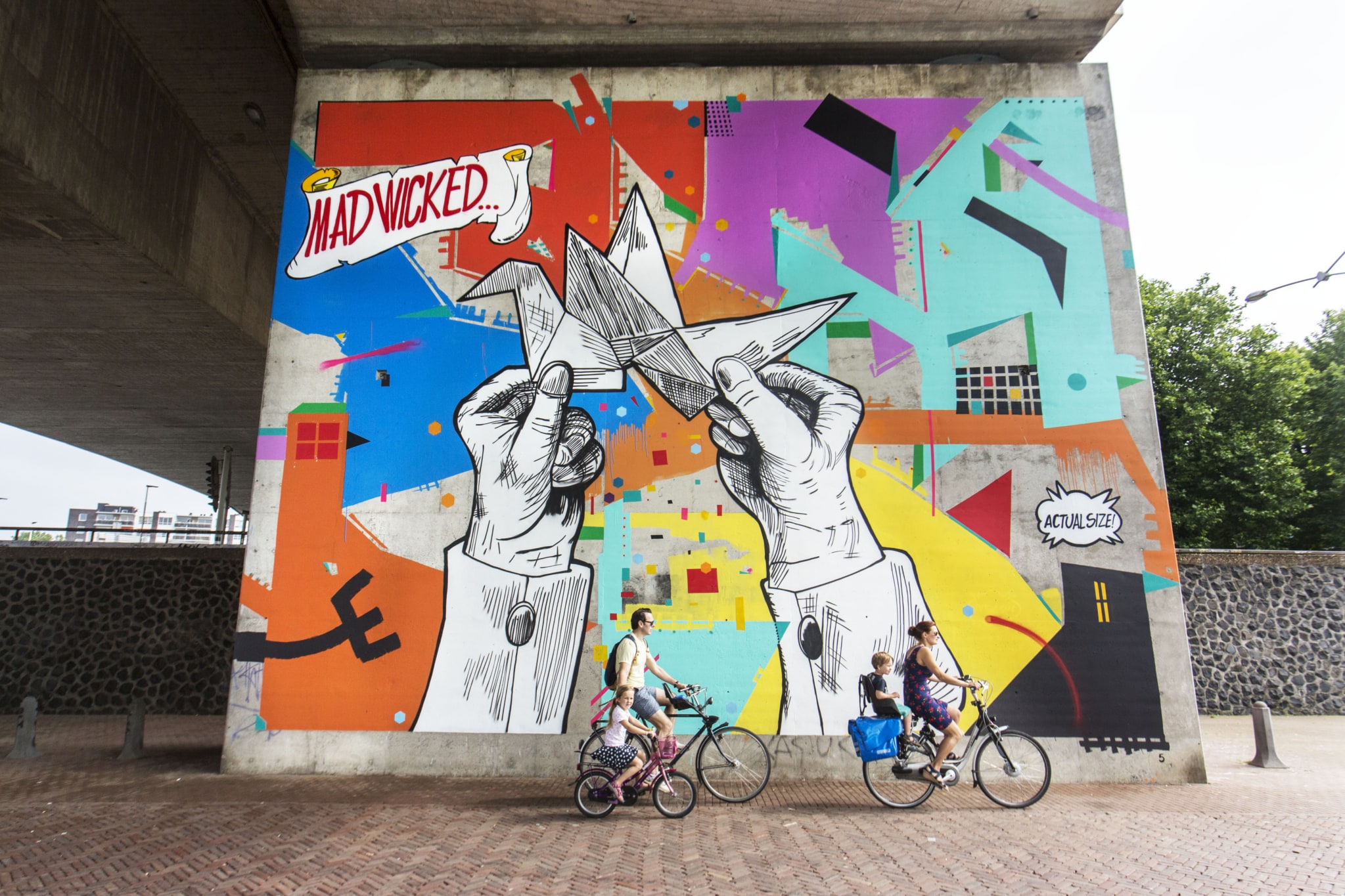 A large mural on a square section of a bridge pillar. The mural stands out with its bright color blocks and large white hands holding up an origami crane. In the top left corner, Mad Wicked is written in red, followed by three dots. In the bottom right corner, in a small speech bubble, are the words actual size. Several sections of the pillar’s concrete have been left unpainted.
