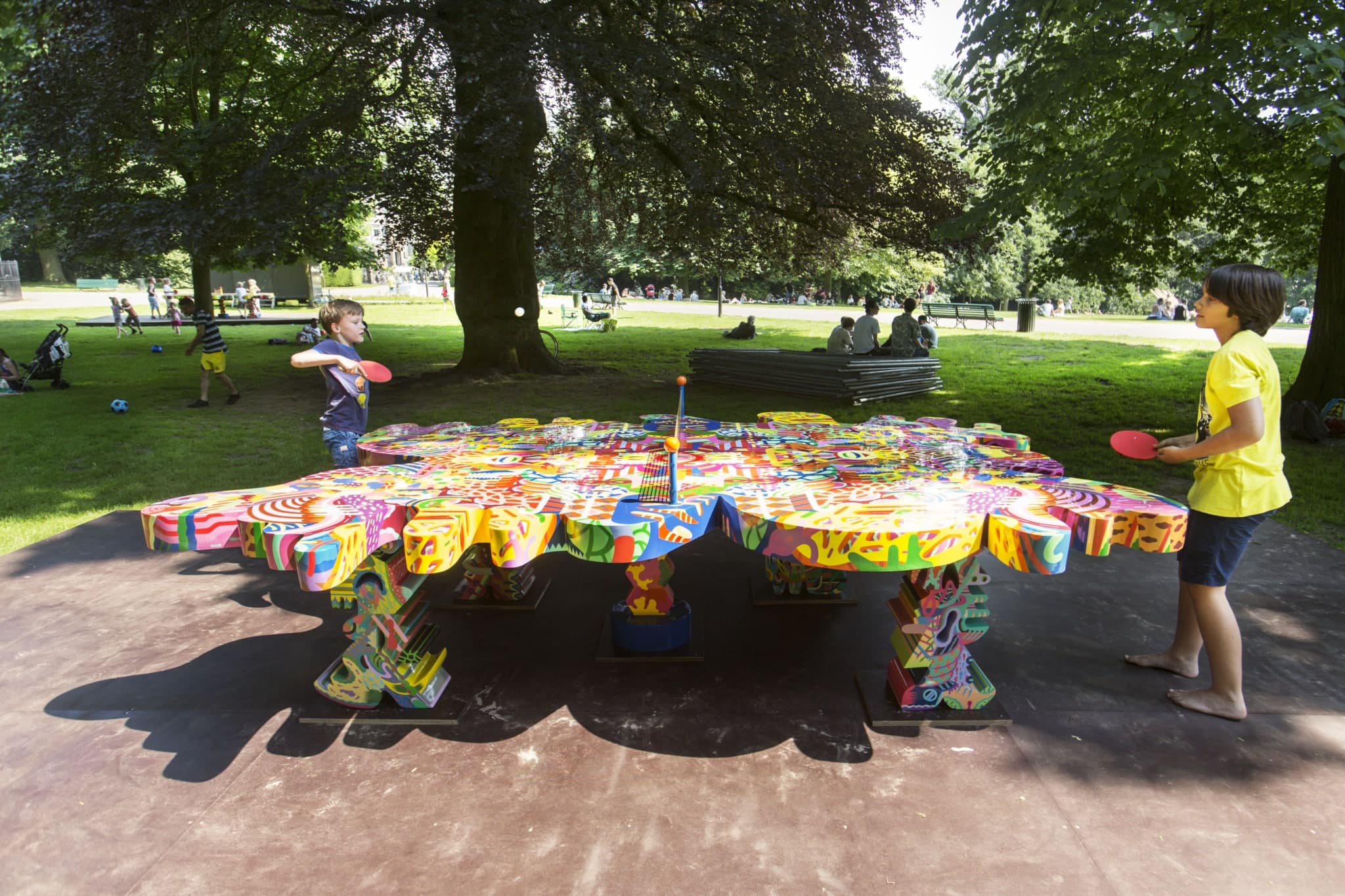 Two children, around ten years old, are playing table tennis on an unusual ping-pong table. The table has an irregular, cloud-like shape. Furthermore, it is not green with white lines but features a vibrant pattern with various abstract shapes and bright colors. A net stands in the middle. The ping-pong table is in the park, where other people are sitting on the grass and children are playing soccer.